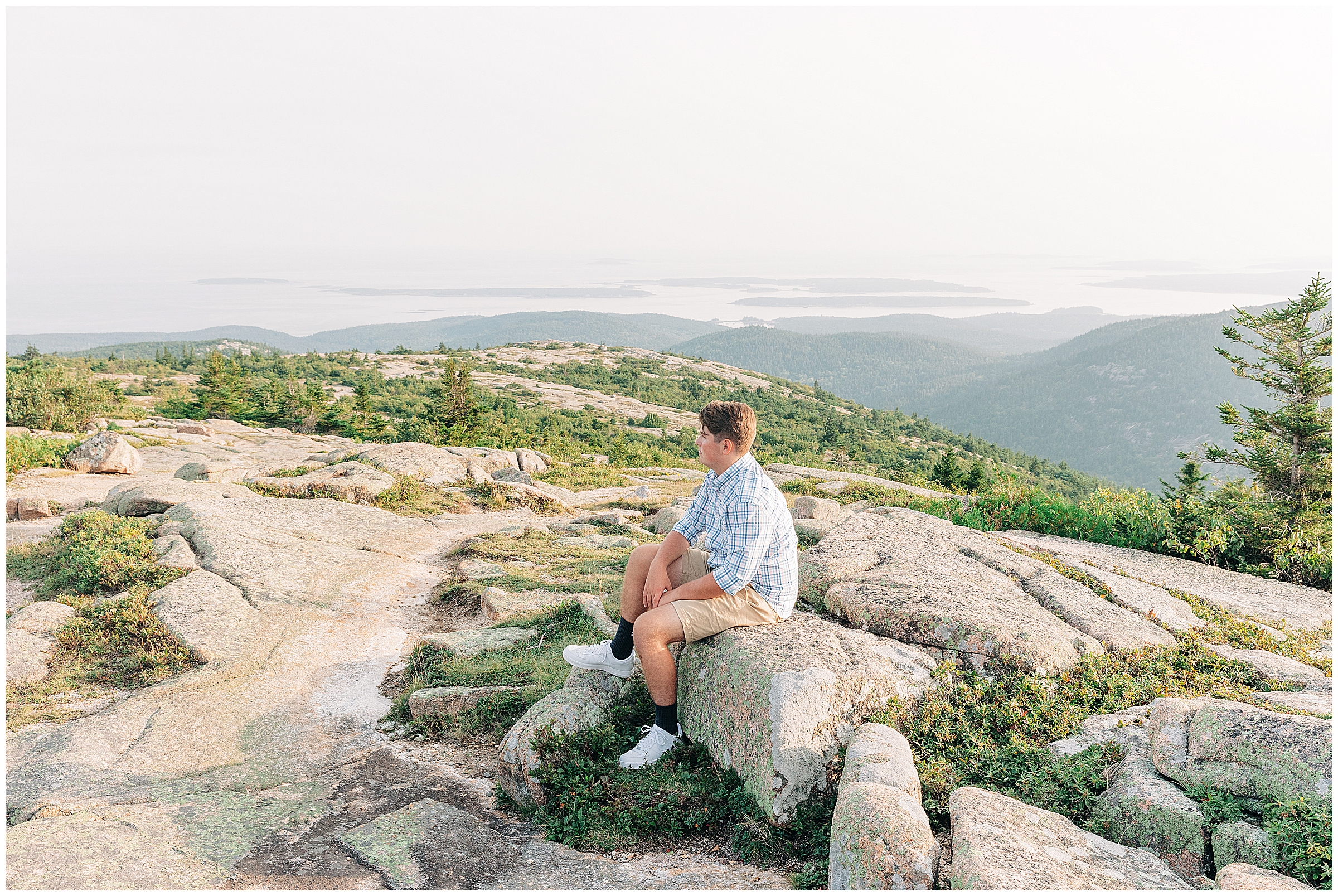 A teenager sits on a rock on top of Cadillac Mountain with the water and islands in the distance